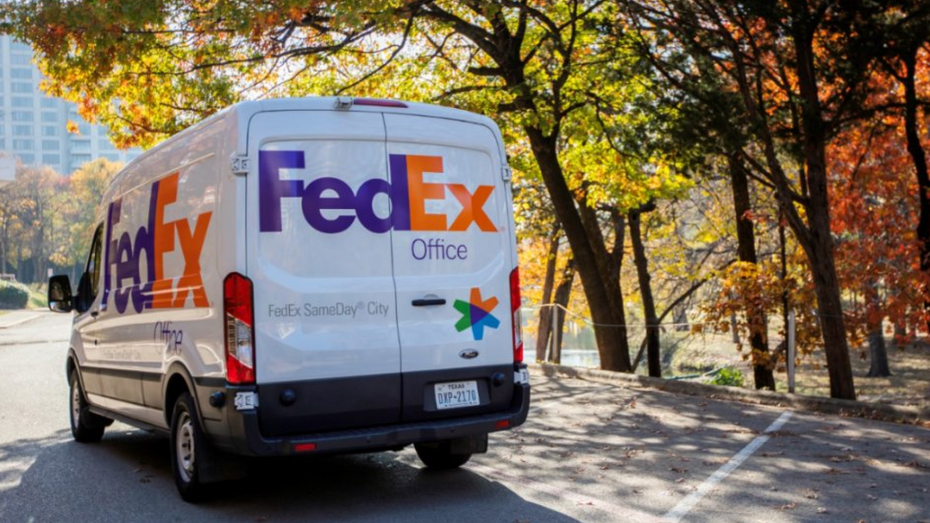 A FedEx SameDay City delivery van is parked under some trees. In the distance are high rise buildings. 