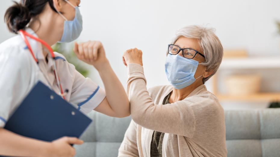 A medical professional with white scrubs, a blue surgical mask, and a blue clipboard elbow-bumps an older Caucasian patient wearing a blue surgical mask and beige cardigan.