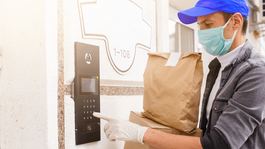 A white delivery man wearing a cloth mask, latex gloves, and cap pushes an apartment call button. He is holding brown paper packages. 