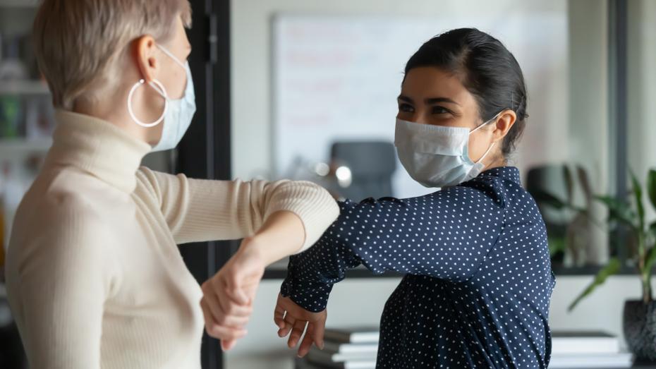 A woman with brown skin and dark hair wearing a navy blouse and blue surgical mask shares a friendly elbow-bump with an older caucasian woman wearing a cream turtleneck, hoop earrings, and a surgical mask. 