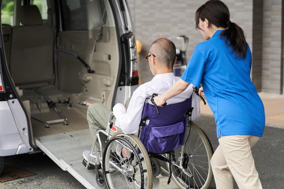 A white woman caregiver pushes the wheelchair of a white man up the ramp of a wheelchair-accessible van.
