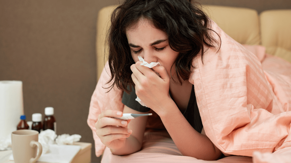 A white woman with rumpled brown hair lies on her stomach under a pink quilt, looking down at a digital thermometer. She holds a tissue to her nose and over her mouth. On her bedside table is a mug, several medicine bottles, and crumpled tissues. 
