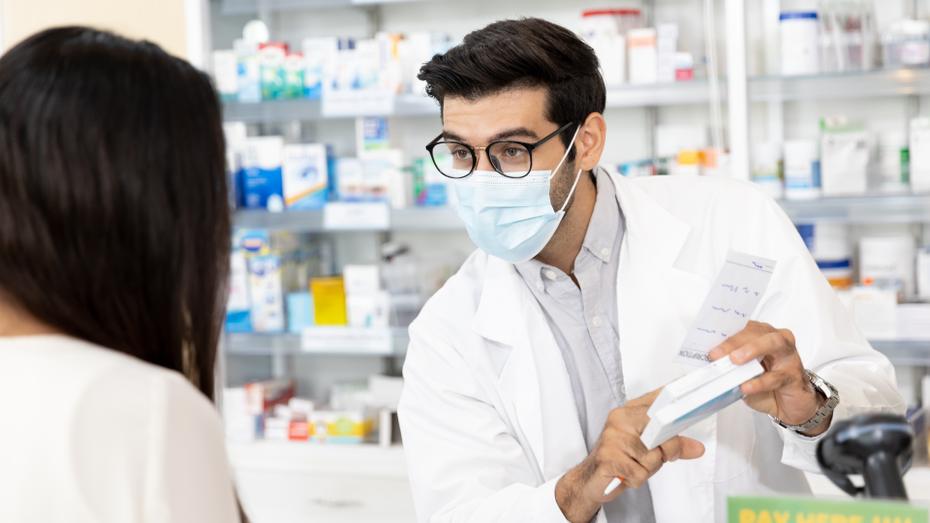 A male pharmacist, wearing glasses, a blue surgical mask, and a white lab coat, explains a prescription to a patient with long dark hair.