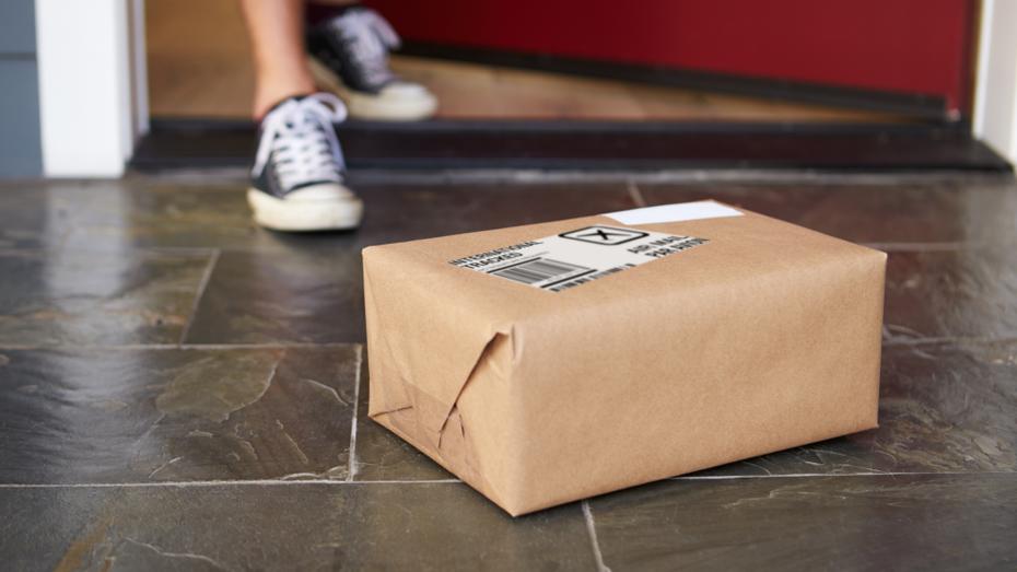 A box wrapped in brown paper with a shipping label on the exterior sits outside a person's front door. The person's feet are in the background as they approach.