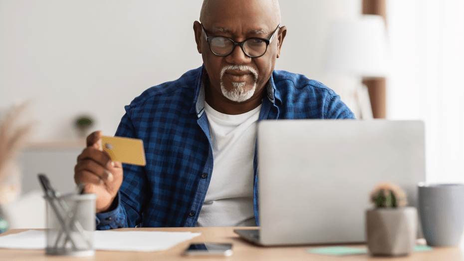 An older black man sits at a desk with a laptop in front of him. He is holding a credit card in his right hand and appears to be using it to pay for something online.