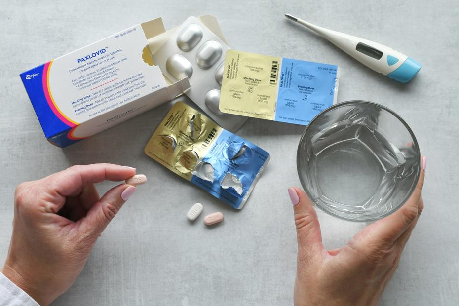 A top-down view of an open box of Paxlovid sitting on a gray table. The morning/night dose packets are visible. A pair of Caucasian hands with painted nails holds a pink nirmatrelvir tablet in one hand and a glass of water in another. 