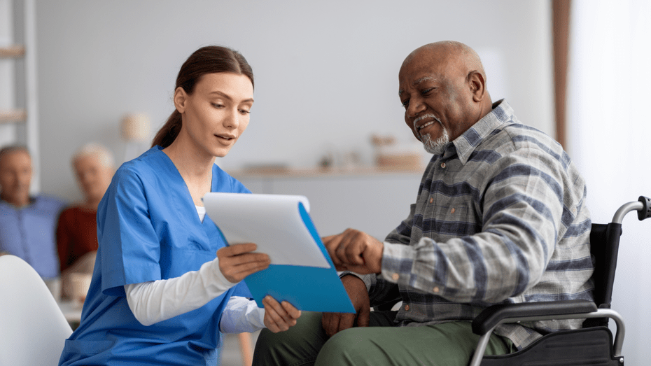 On the left, a white woman in blue scrubs angles a blue clipboard toward the patient next to her. On the right, the patient, an older Black man in a plaid shirt and using a wheelchair, smiles as he reads the clipboard.