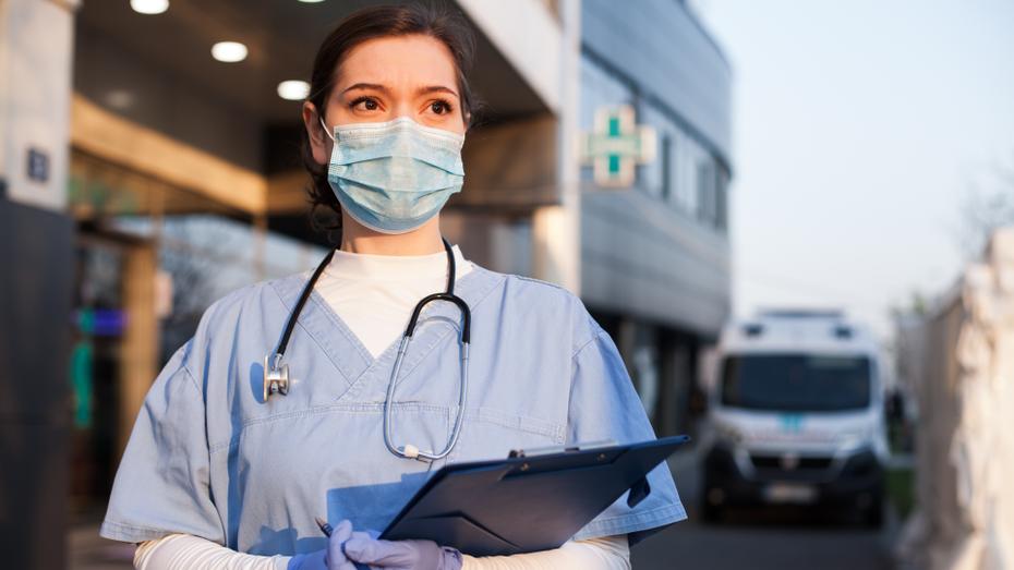 A woman wearing a surgical mask, gloves, and scrubs with a clipboard in her hands and a stethoscope around her neck looks into the distance.