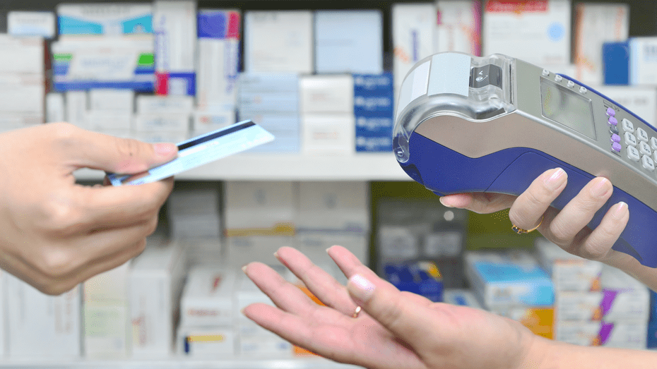 In a pharmacy setting, a person hands a credit card to another person who is holding a card reader. 