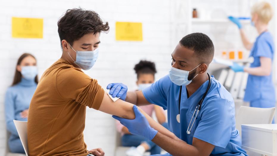 A person in a brown shirt receives the COVID-19 vaccine from a healthcare professional wearing blue scrubs.